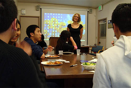Student leadership workshop at UC San Diego - students sit around a table eating pizza in foreground while a standing facilitator talks with the group